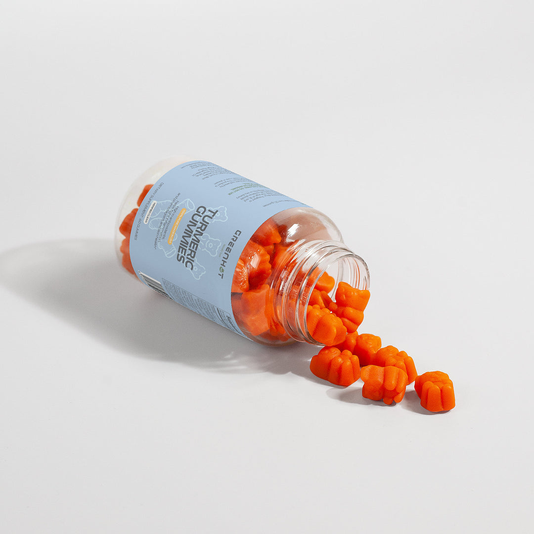 A plastic bottle with a light blue label lies on its side, spilling out GreenHat Turmeric Gummies - Immunity Accelerator shaped like small bears onto a white surface.