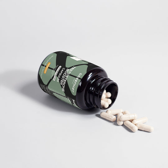 A bottle of dietary supplement capsules lies on its side with some white capsules spilled out onto a white surface. The bottle, labeled as a Testosterone Booster - Get The Most Out Of Yourself, features a green and black design by GreenHat.