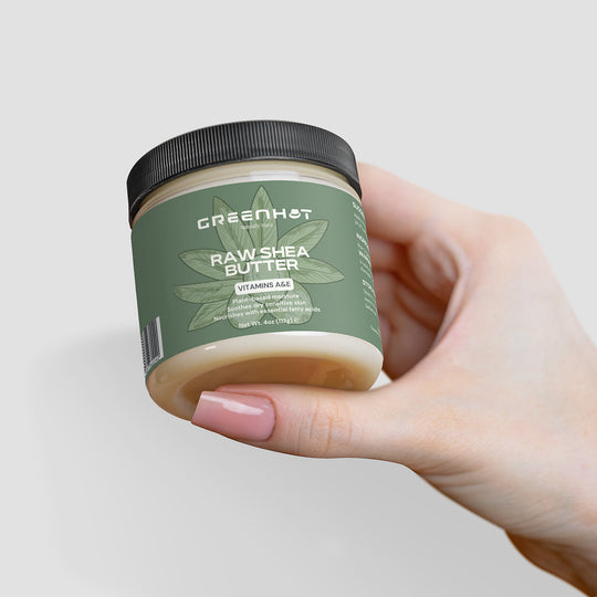 A hand holding a jar of GreenHat Raw Shea Butter. The jar has a green label with text indicating its unrefined goodness and volume (113g/4oz). The person's nails are painted light pink.