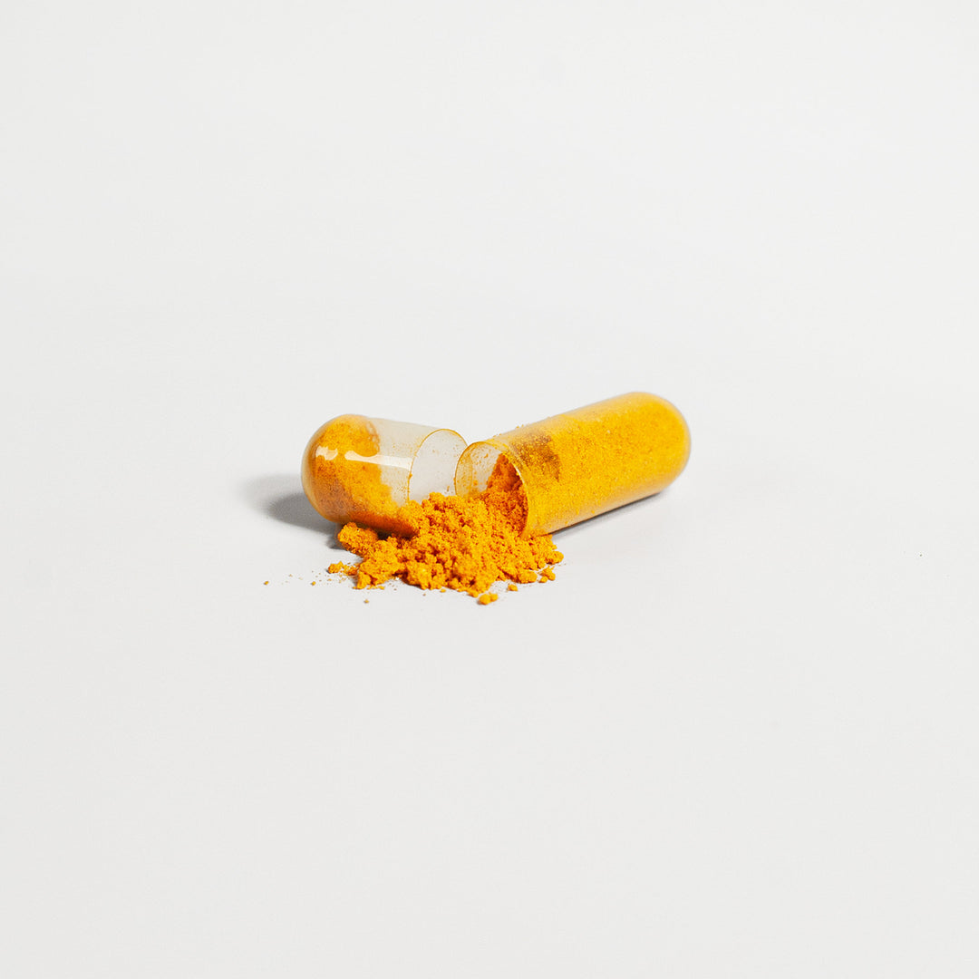 A yellow powder, indicative of a Platinum Turmeric - Immunity Accelerator supplement from GreenHat, is spilling out of a transparent capsule split in half against a white background.