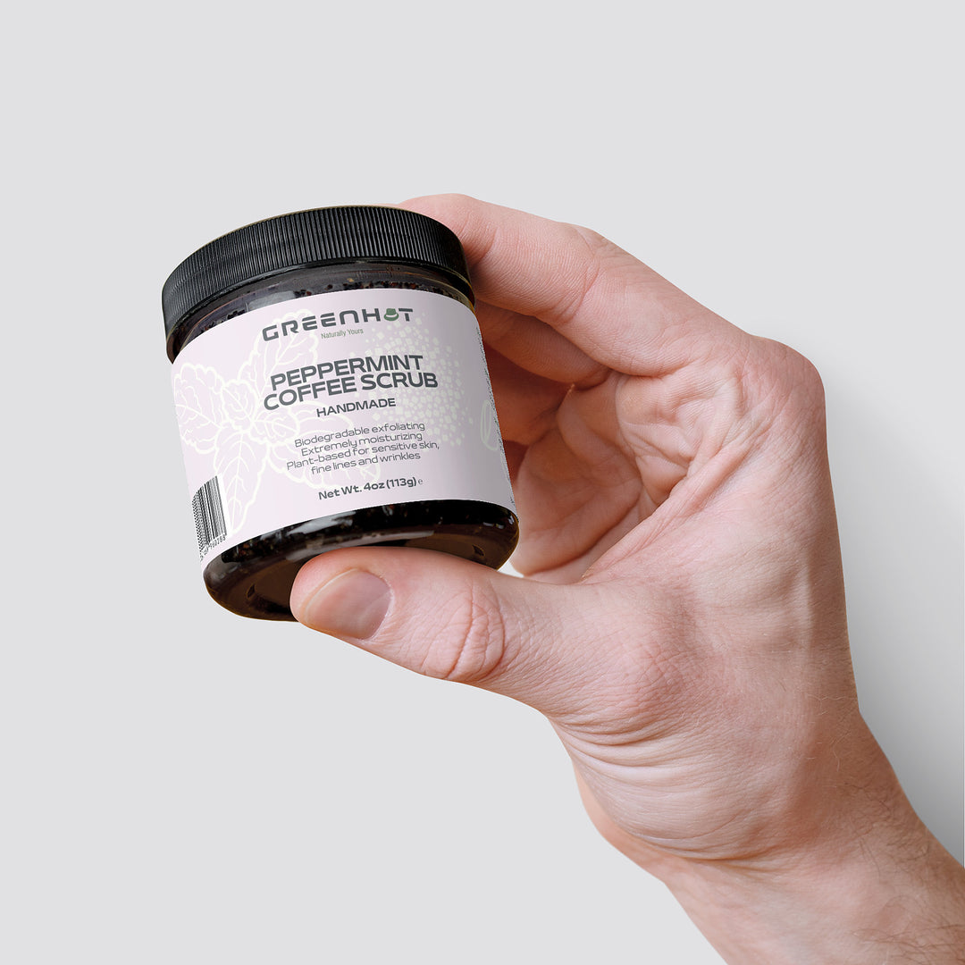 A hand holding a jar of "GreenHat Peppermint Coffee Scrub," an antioxidant-rich coffee scrub with a white label detailing ingredients and product weight of 113g (4oz), against a plain background.