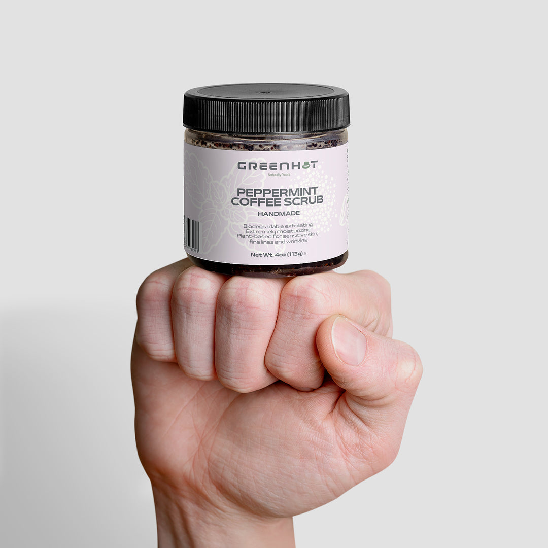 A hand holding a jar of GreenHat Peppermint Coffee Scrub, labeled as handmade and weighing 4 oz (113g). This antioxidant-rich coffee blend promises effective exfoliation.