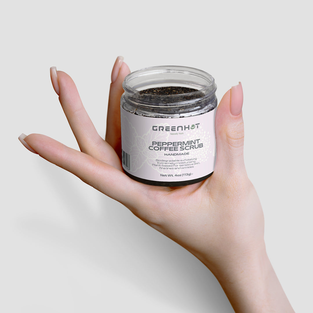 A hand is holding a small jar of GreenHat Peppermint Coffee Scrub, known for its antioxidant-rich coffee and effective exfoliation, with a white label.