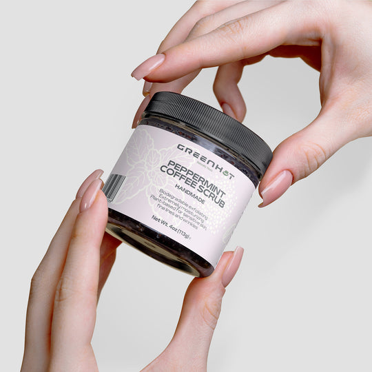 Hands hold a jar of GreenHat's Peppermint Coffee Scrub, known for its antioxidant-rich coffee and effective exfoliation, with a black lid, white label, and floral design, against a white background.