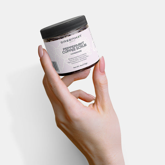 A hand holding a jar of GreenHat Peppermint Coffee Scrub, known for its effective exfoliation and antioxidant-rich coffee, is shown against a white background.