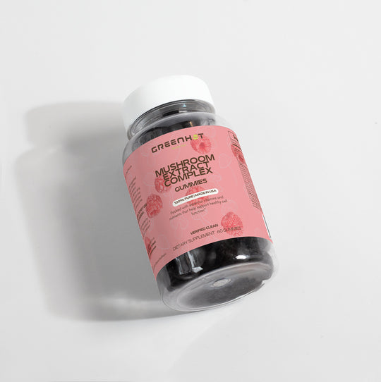 A bottle of GreenHat Mushroom Extract Complex Gummies for stress relief and immunity boost, with a pink label, lying on its side on a white surface.