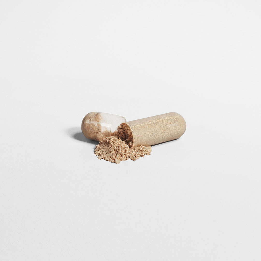 A transparent capsule with beige powder inside, partially opened, with some GreenHat Mushroom Complex 10 X - Natural Power supplements spilled out on a plain white background.