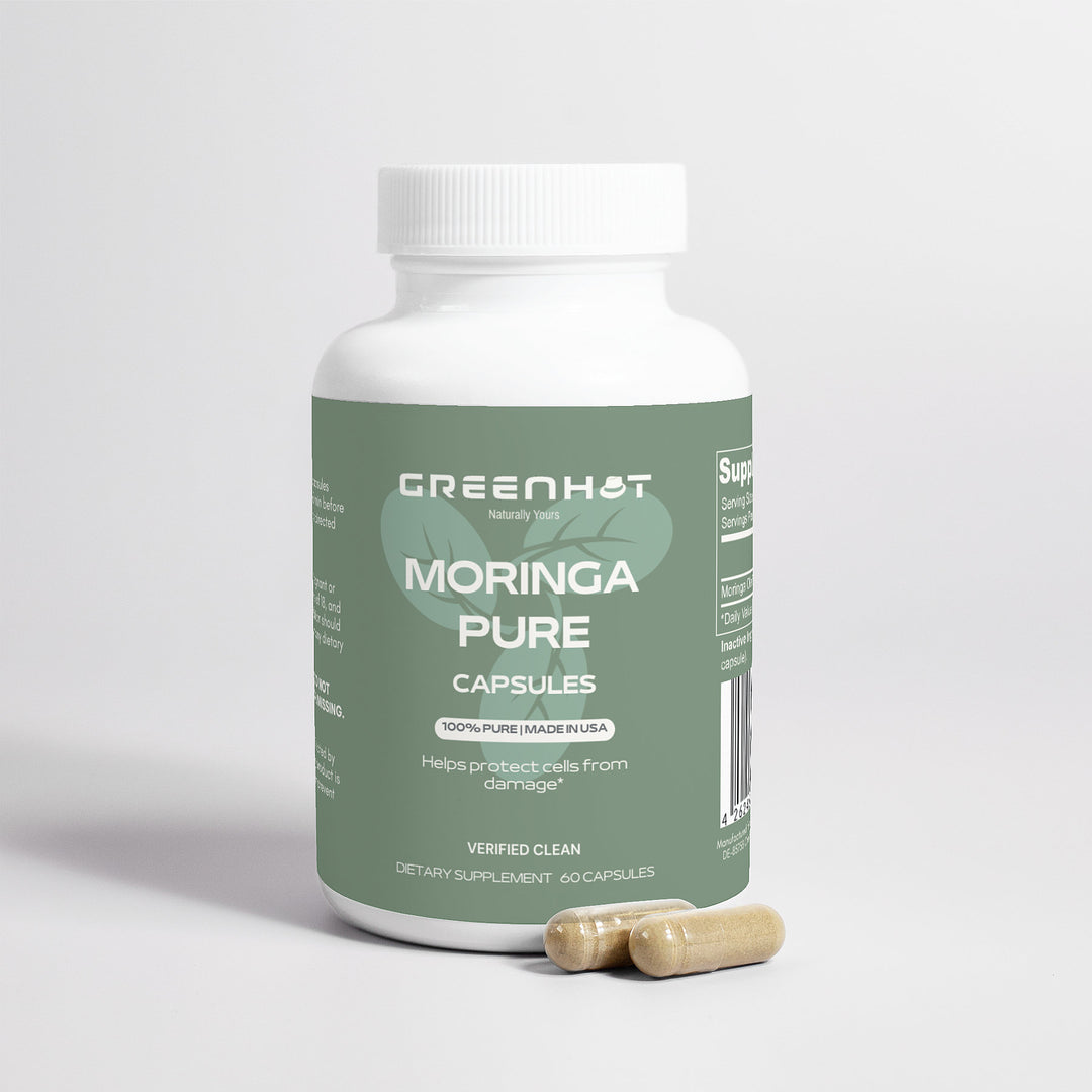 A bottle of GreenHat Moringa Pure - Enhance Cellular Health capsules stands on a white surface, with two capsules placed beside it. The label indicates this superfood helps protect cells from damage and is made in the USA.