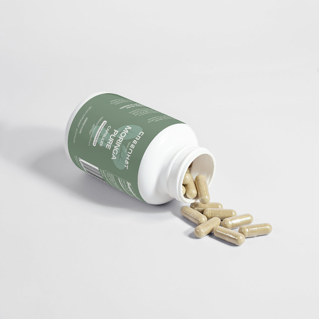 A green bottle of Moringa Pure - Enhance Cellular Health herbal capsules lies on its side with capsules spilling out on a white surface. The label reads "GreenHat." This superfood supplement is designed to elevate your daily wellness routine.