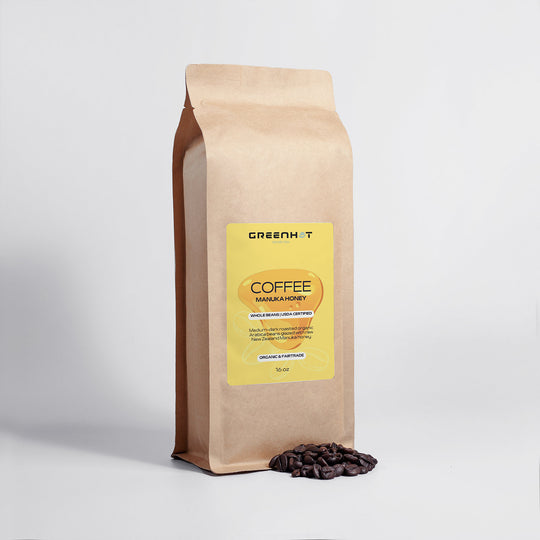 A brown bag of GreenHat-brand coffee sits upright against a white background, with Arabica beans scattered near its base. The label reads "Manuka Honey Coffee 16oz - Awaken Your Senses" with additional details below, hinting at the rich flavors inspired by New Zealand's finest.