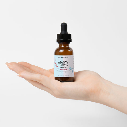 A hand holds a small brown bottle with a dropper labeled "Scalp Health & Hair Growth Hair Oil" by GreenHat, made with natural oils for promoting healthy hair growth.