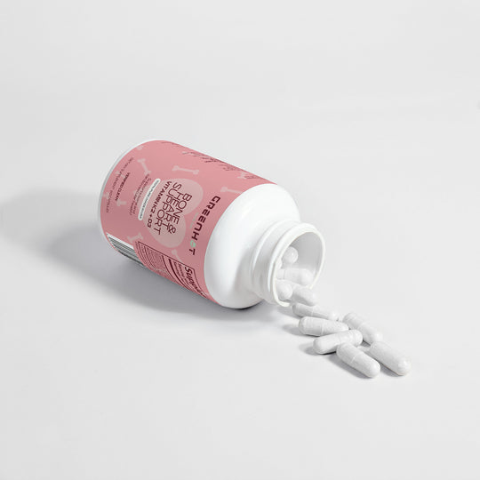 White capsule pills spilling out of an open white bottle with a pink label, promoting bone support. The bottle, lying on a pristine white surface, highlights the benefits of Vitamin K2 and Vitamin D3 for overall health from GreenHat's Bone & Heart Support - Strengthen Your Foundation.