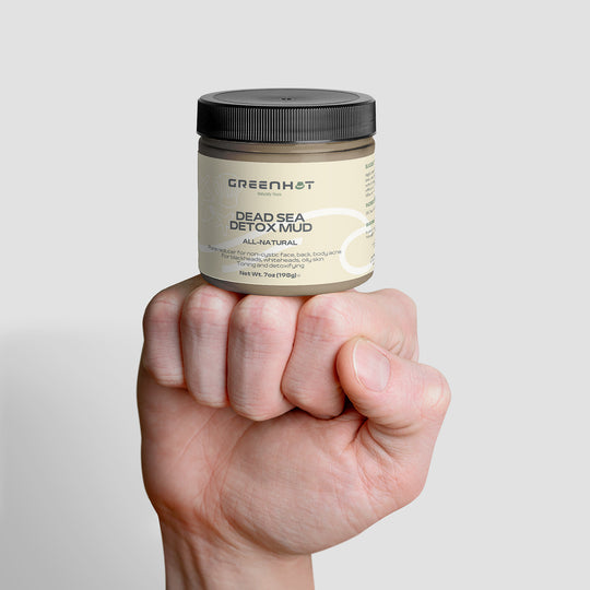 Hand holding a jar of GreenHat Dead Sea Detox Mud for skin rejuvenation against a plain background.