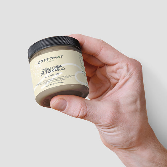 A hand holding a jar of natural GreenHat Dead Sea Detox Mud for skin rejuvenation against a white background.