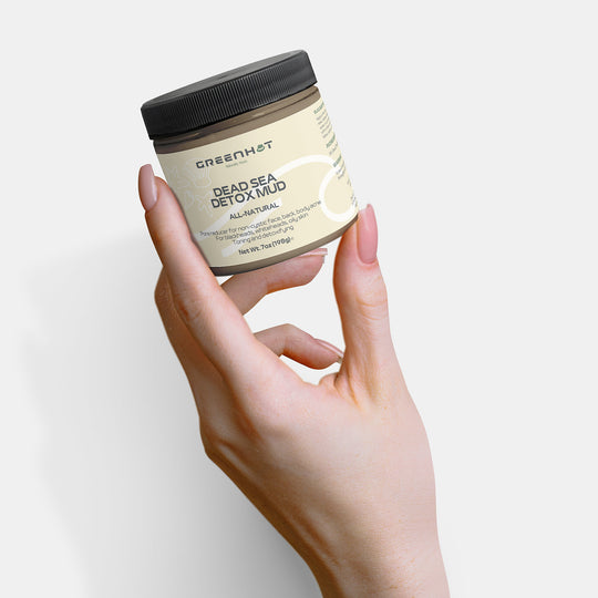 A hand holds a jar of GreenHat Dead Sea Detox Mud for skin rejuvenation against a white background.