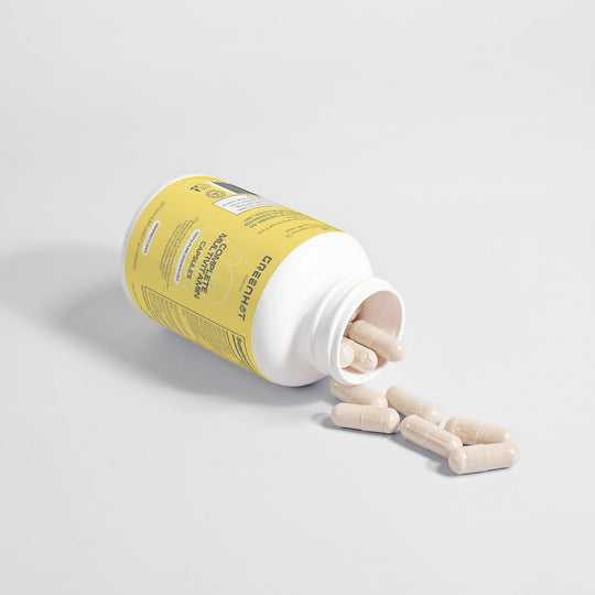 A white bottle with a yellow label is tipped over, spilling GreenHat Complete Multivitamin capsules on a light gray surface.