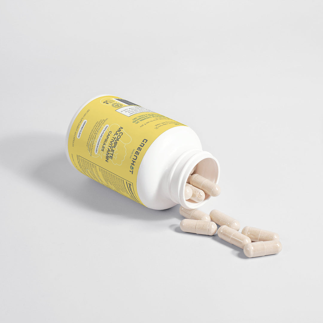 A plastic bottle labeled "GreenHat Complete Multivitamin" with a yellow label is tipped over on a white surface, with beige capsules spilling out. These multivitamin capsules combine immune-boosting herbs and antioxidants for optimal health support.