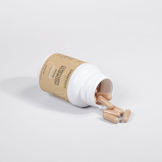 A white plastic bottle with a beige label, containing beige capsules of Cordyceps Mushroom - Enhanced Physical Performance by GreenHat, is tipped over with some capsules spilling out.