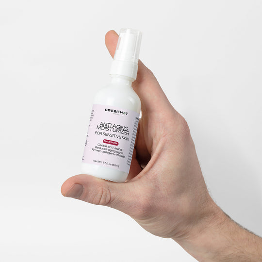 A person's hand holding a bottle of GreenHat Anti Aging Moisturizer for Sensitive Skin against a white background.