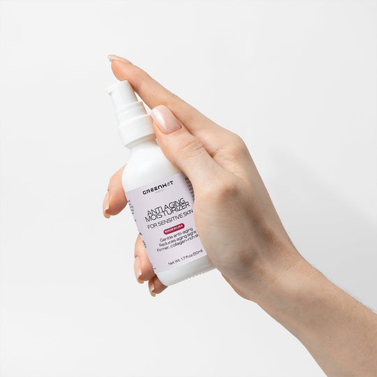 A hand holding a white bottle of GreenHat Anti Aging Moisturizer for Sensitive Skin with a pump dispenser against a plain white background.