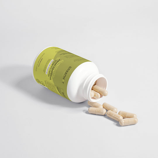 A plastic bottle with a green label lies on its side, spilling beige capsules of GreenHat Ashwagandha - Unlock Your Inner Energy onto a white surface.