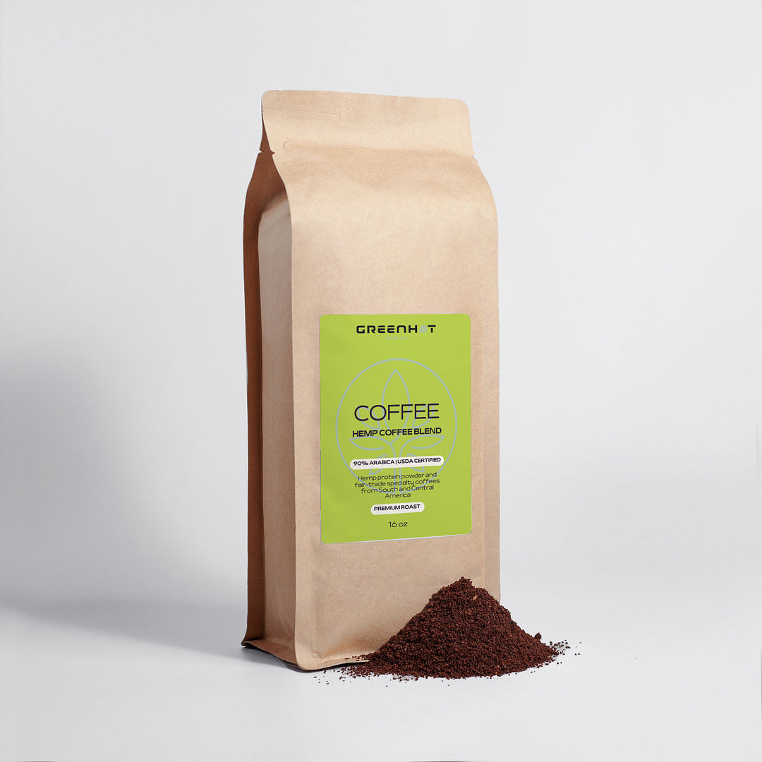 A beige bag of GreenHat Organic Hemp Coffee Blend - Medium Roast 16oz sits upright with a small pile of ground coffee at its base. The green label highlights antioxidants, serving size, and the benefits of hemp protein powder.