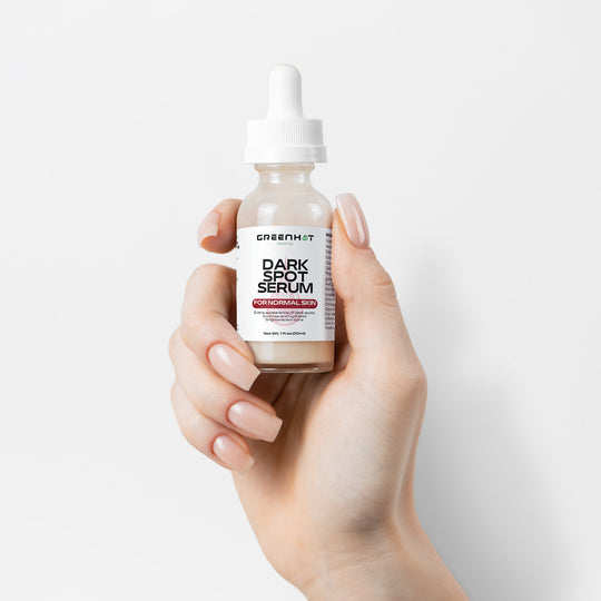 A hand holding a small bottle of GreenHat Dark Spot Serum for Normal Skin with a dropper cap and a label that reads "Paraben Free," ideal for achieving an even skin tone.