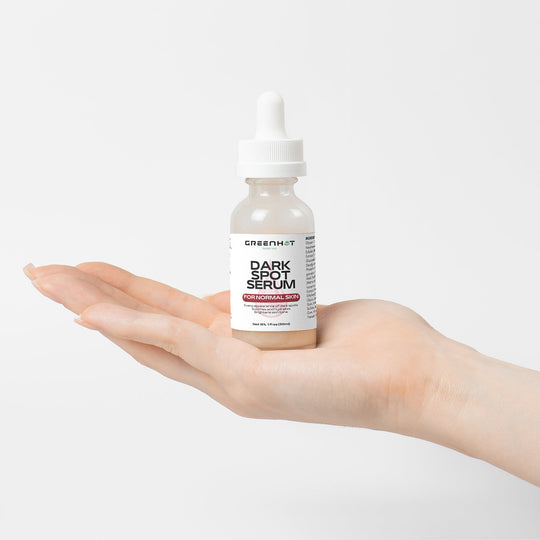 A hand holds a bottle labeled "Dark Spot Serum for Normal Skin" with a dropper cap, by the brand GreenHat, promising dark spot reduction and an even skin tone, against a neutral background.