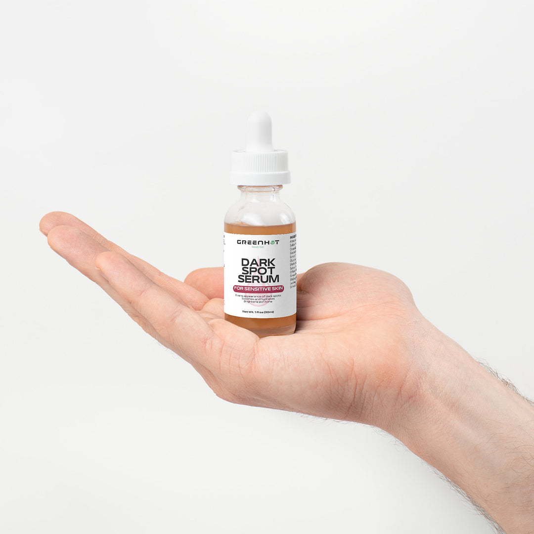 Hand holding a bottle of GreenHat Dark Spot Serum for Sensitive Skin with a dropper top and white label on a white background. Formulated with natural extracts, this serum is gentle enough for sensitive skin.