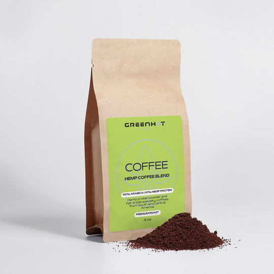 A brown paper bag labeled "GreenHat Organic Hemp Coffee Blend - Medium Roast 4oz" sits upright with ground coffee spread at its base on a plain white background, highlighting the blend's commitment to Fair Trade Coffee.