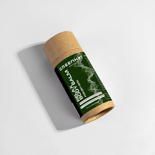 A cardboard tube labeled "GreenHat Relax Body Balm" lays on a white surface, casting a shadow, offering both moisturizing treatment and aromatherapy benefits.