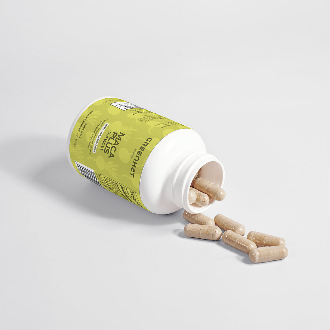 A bottle of Maca Plus - Experience Natural Vitality dietary supplement capsules is shown with the lid open and several tan capsules spilled out onto a white surface. The bottle, promoting an energy boost, has a green GreenHat label with text and branding.