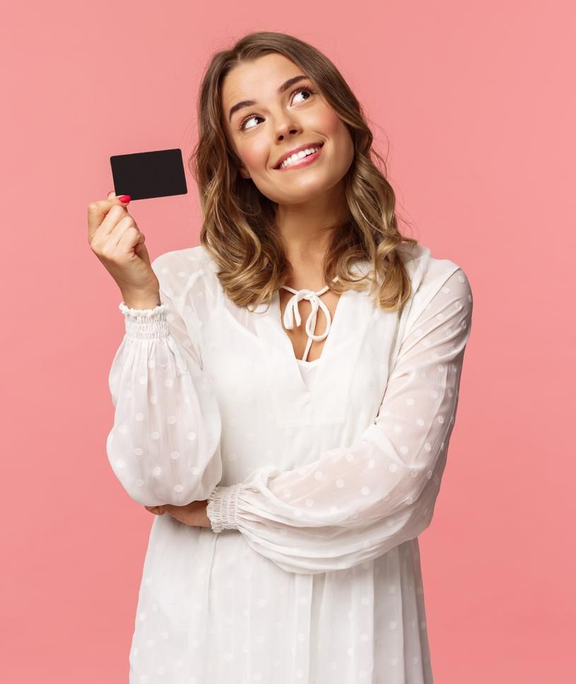 A person in a white dress holds a GREENHAT Gift Voucher by GreenHat while smiling and looking upwards against a pink background.