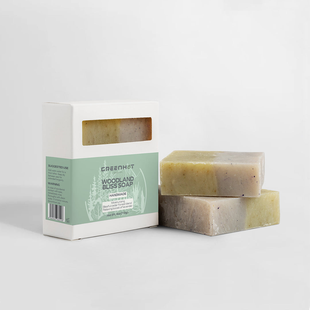 Two bars of GreenHat's "Woodland Bliss" soap beside their packaging on a white background. The box displays product ingredients and branding.