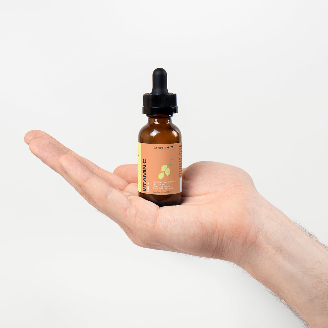A person's hand holding a brown glass dropper bottle labeled "GreenHat Vitamin C Serum - Hydration and Skin Tone Enhancement" against a white background.
