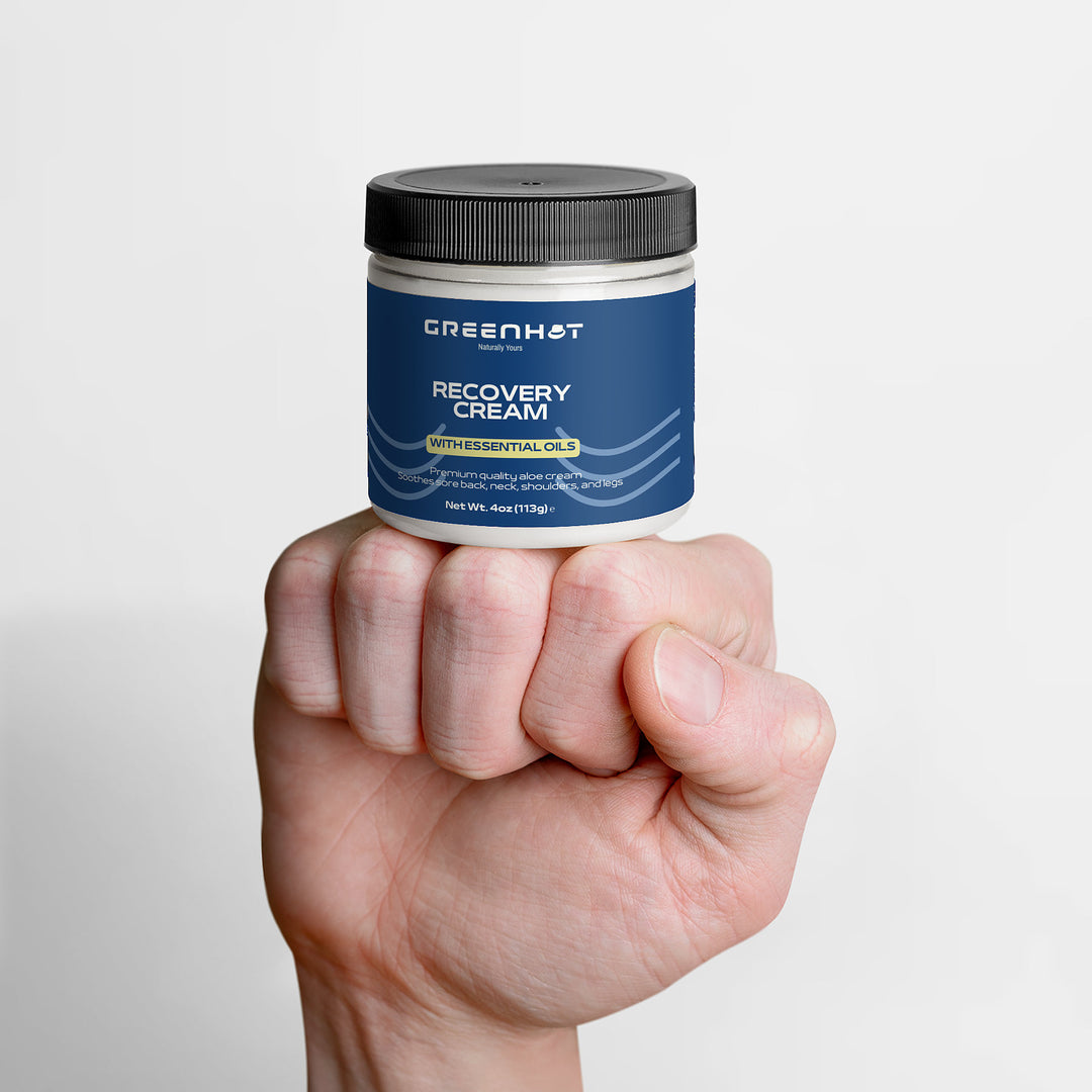 A hand holding a small jar of GreenHat premium recovery cream against a white background.