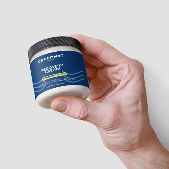 A hand holding a jar of GreenHat premium cosmetic recovery cream with essential oils against a white background.