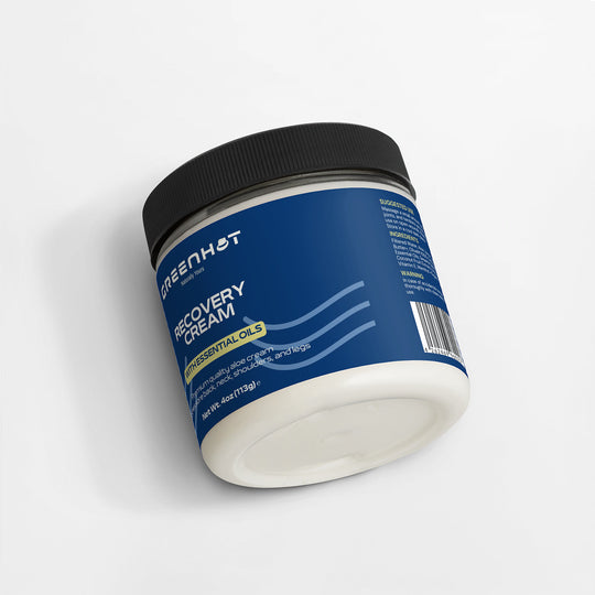 A blue-labeled jar of GreenHat premium Recovery Cream with essential oils resting on a white surface, viewed from a slight angle emphasizing the label.