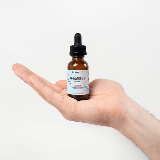 A caucasian hand holding a dropper bottle labeled as "GreenHat Moisturizing and Strengthening Hair Oil with added sweet almond oil" against a white background.