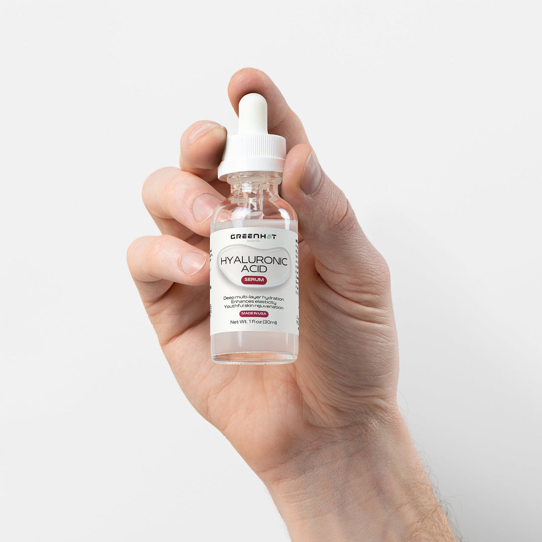 A hand holding a small bottle of GreenHat Hyaluronic Acid Serum for multi-layer hydration with a dropper, isolated on a white background.