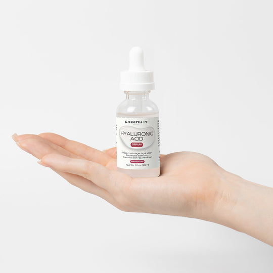 A hand holding a bottle of GreenHat Hyaluronic Acid Serum, designed for multi-layer hydration, against a plain white background.
